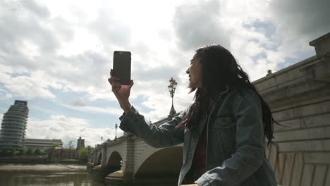 hispanic latina tourist using skype and showing her friend the river thames and putney bridge in london, smiling and having a great time