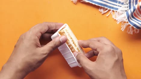 hands holding a wafer sandwich cookie in a plastic container on an orange surface with a striped napkin