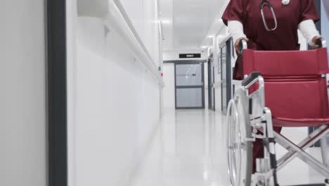 african american female doctor running with wheelchair in hospital corridor, slow motion