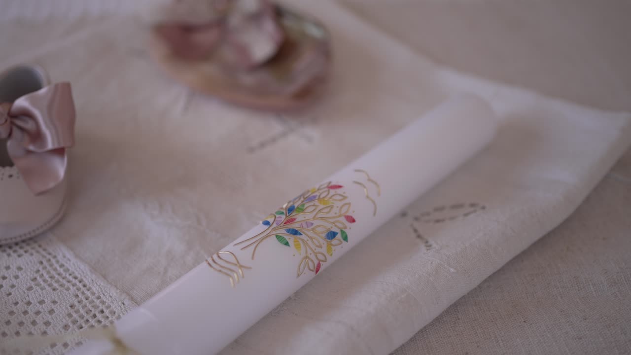 vela de bautismo decorativa con diseño floral, emparejada con zapatos de bebé y una concha de mar, colocada en una tela cubierta de encaje, que simboliza una ceremonia sagrada