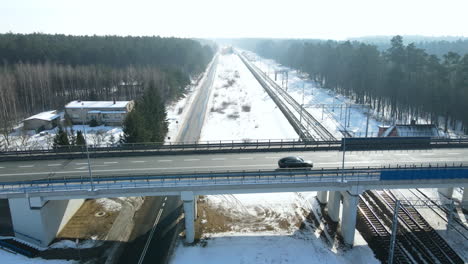 Autos,-Die-über-Ein-Viadukt-Fahren,-Das-Eine-Andere-Schwarze-Straße-Und-Drei-Gleise-Einer-Eisenbahn-In-Der-Nähe-Eines-Bahnhofs-Zwischen-Den-Bäumen-In-Einer-Winterlandschaft-überquert