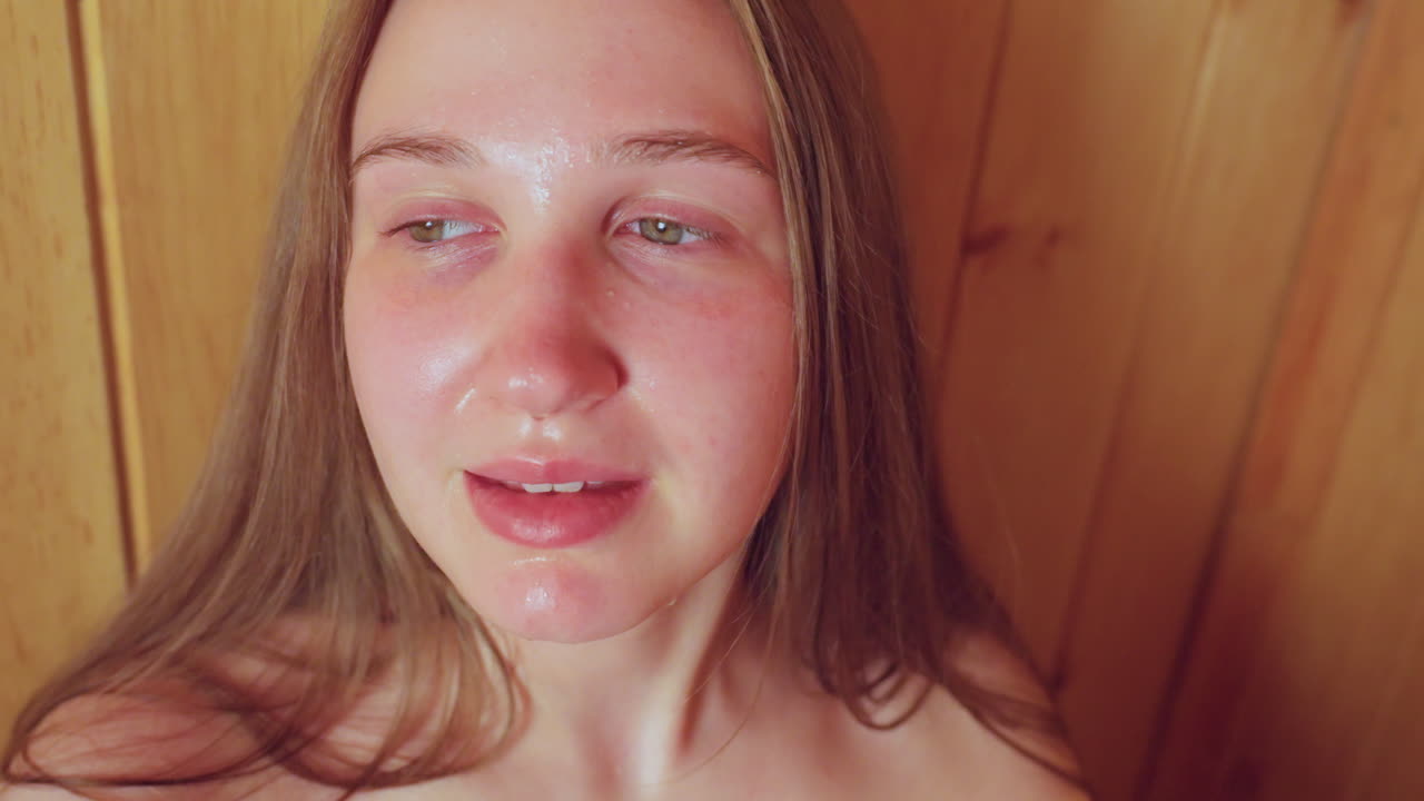 lose up of young woman with long hair holding warm cup of tea with both hands inside cozy wooden sauna, eyes closed in relaxation, soft smile as she inhales aroma of soothing herbal infusion