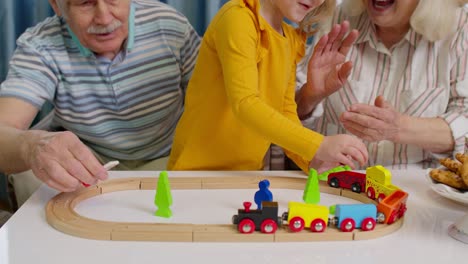 senior grandparents with child kid granddaughter playing game, riding toy train on railway at home