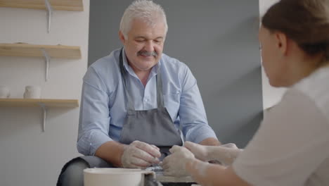 A-woman-master-shares-her-skills-with-a-senior-man-conducting-a-lesson-for-the-elderly.-Show-grandfather-the-technique-of-working-on-a-potter's-wheel-with-ceramic-clay