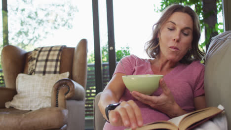 Relaxed-caucasian-pregnant-woman-lying-on-sofa,-eating-and-reading-book