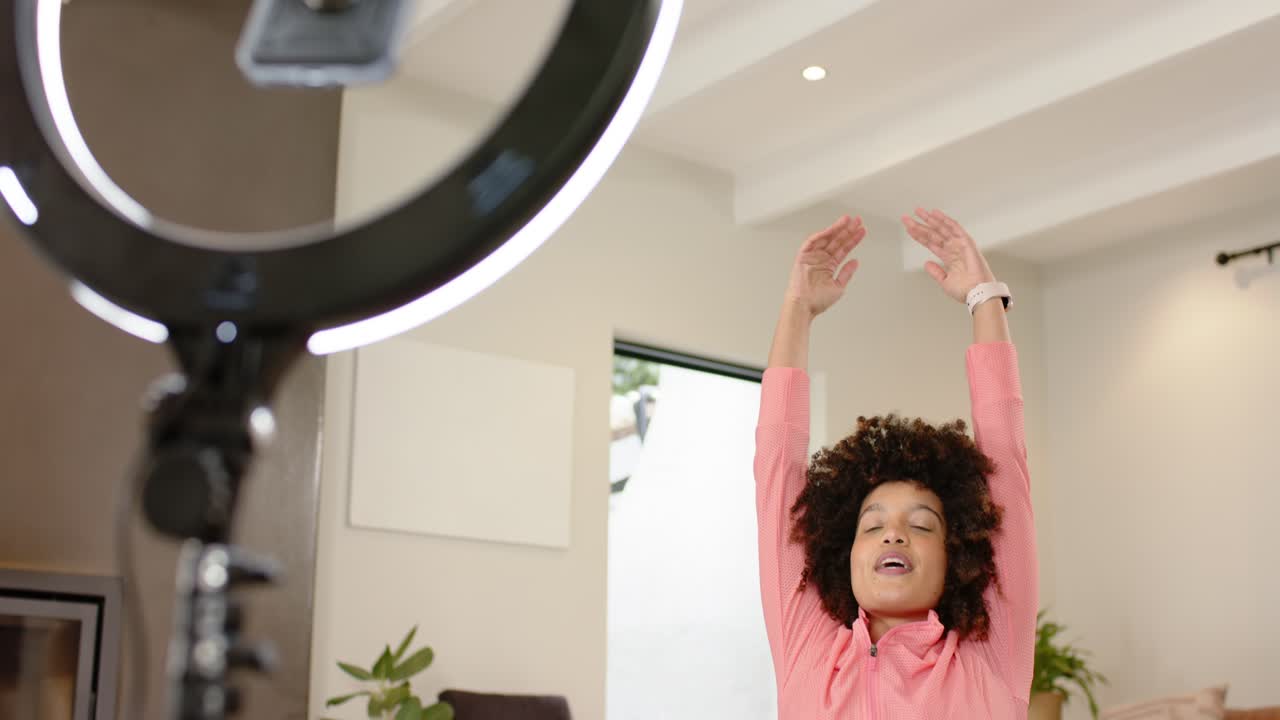 Woman kneeling at home facing ring light, stretching arms overhead, camera zooming for filming