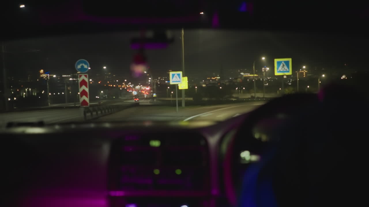 nærmer seg nattlig bysilhuett gjennom frontruten, bud bak rattet navigerer på motorveien mot fjerne lys, dashbordglød og neonrefleksjoner skaper filmatisk bevegelse, urbane rute
