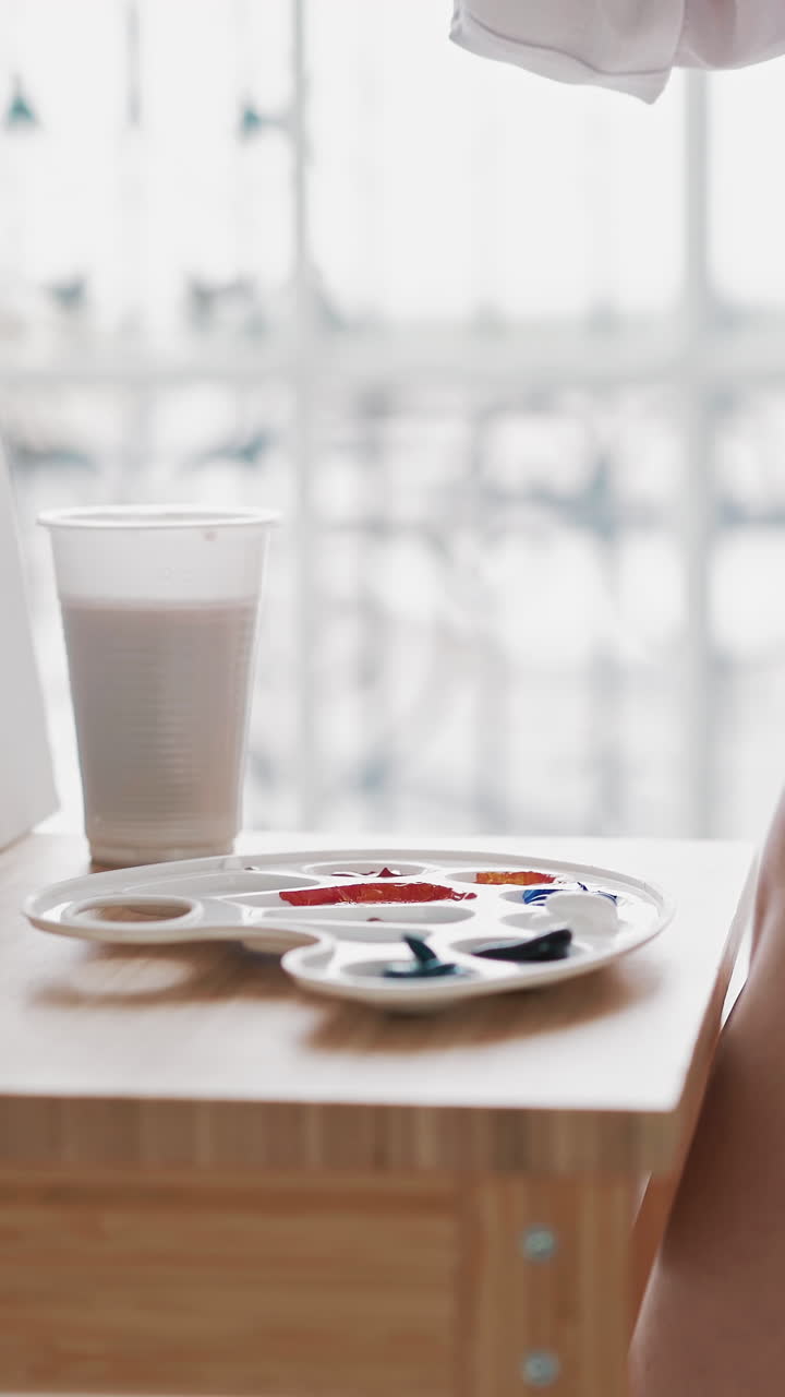 Talented woman artist in stylish clothes draws taking paint from palette with brush sitting near wooden easel with canvas in art studio closeup