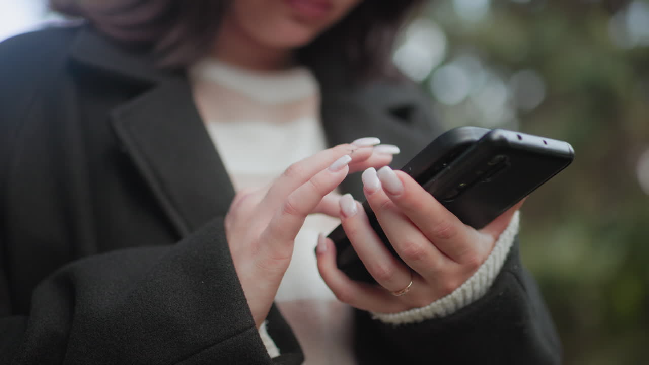 두 개의 휴대폰을 들고 화면을 터치하며 밝은 표정을 짓고 있는 우아한 젊은 여성의 로우 앵글 뷰, 부드러운 보케 배경, 아늑한 코트, 현대적인 소통의 순간