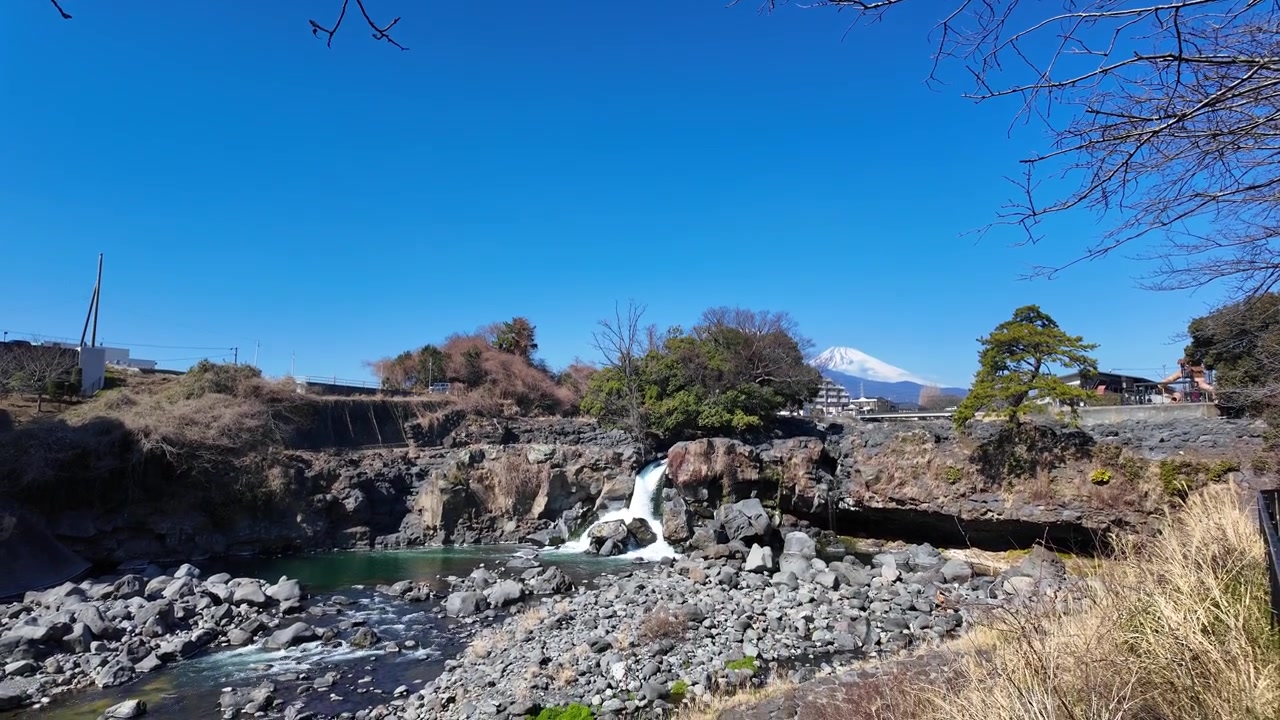 Berrak mavi bir gökyüzü altında, arka planda Fuji Dağı'nın göründüğü şelalenin geniş açılı manzarası.