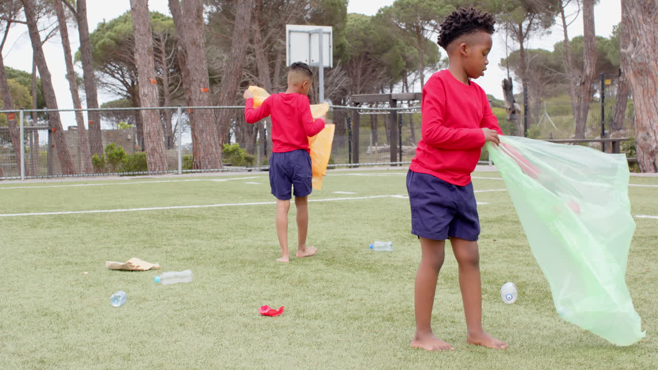 Multiracial boys cleaning up litter on school field, holding colorful trash bags