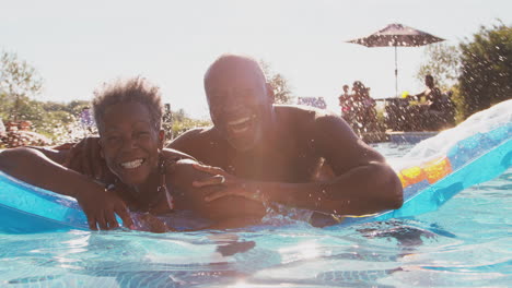 Portrait-Of-Smiling-Senior-Couple-On-Summer-Holiday-Relaxing-In-Swimming-Pool-On-Inflatable-Airbed