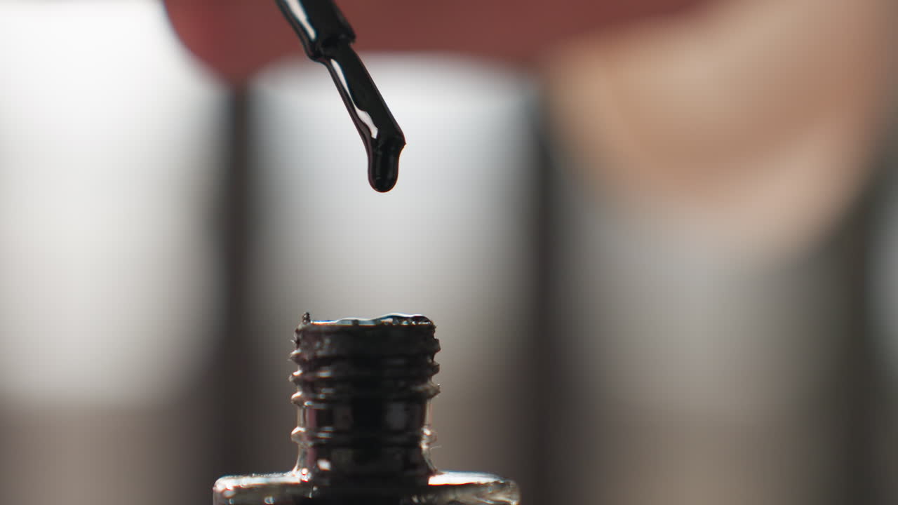 Close up black nail polish drop falls from brush into transparent bottle, glossy liquid bead drips back toward neck, macro beauty detail with shallow depth, blurred background, manicure concept