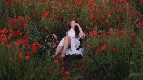 Weite-Aufnahme-Eines-Wunderschönen-Dunkelhaarigen-Mädchens-Mit-Einer-Mohnblume-Im-Haar,-Das-In-Einem-Feld-Aus-Wildblumen-Und-Roten-Mohnblumen-Liegt,-Ein-Kleid-Trägt-Und-Lächelt