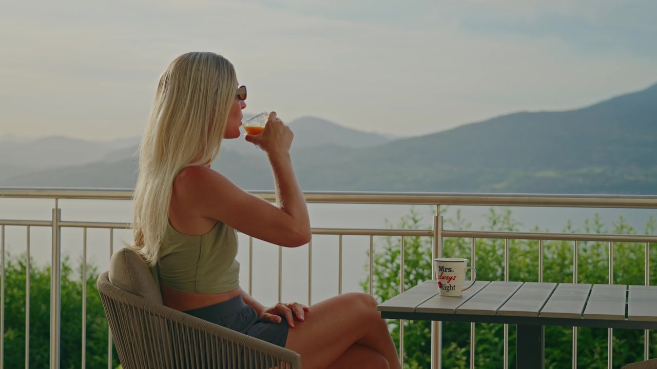 Una donna caucasica che indossa occhiali da sole si gode un succo d'arancia fresco su un balcone con vista sul Lago Maggiore nella soffusa luce del mattino. Splendide viste sulle montagne e sul lago creano una rilassante scena estiva italiana.
