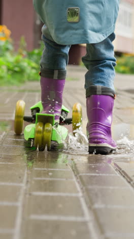 child rides kick scooter on rainy day. playful kid wearing rubber boots enjoys scooter riding on wet pavement close backside view. healthy childhood