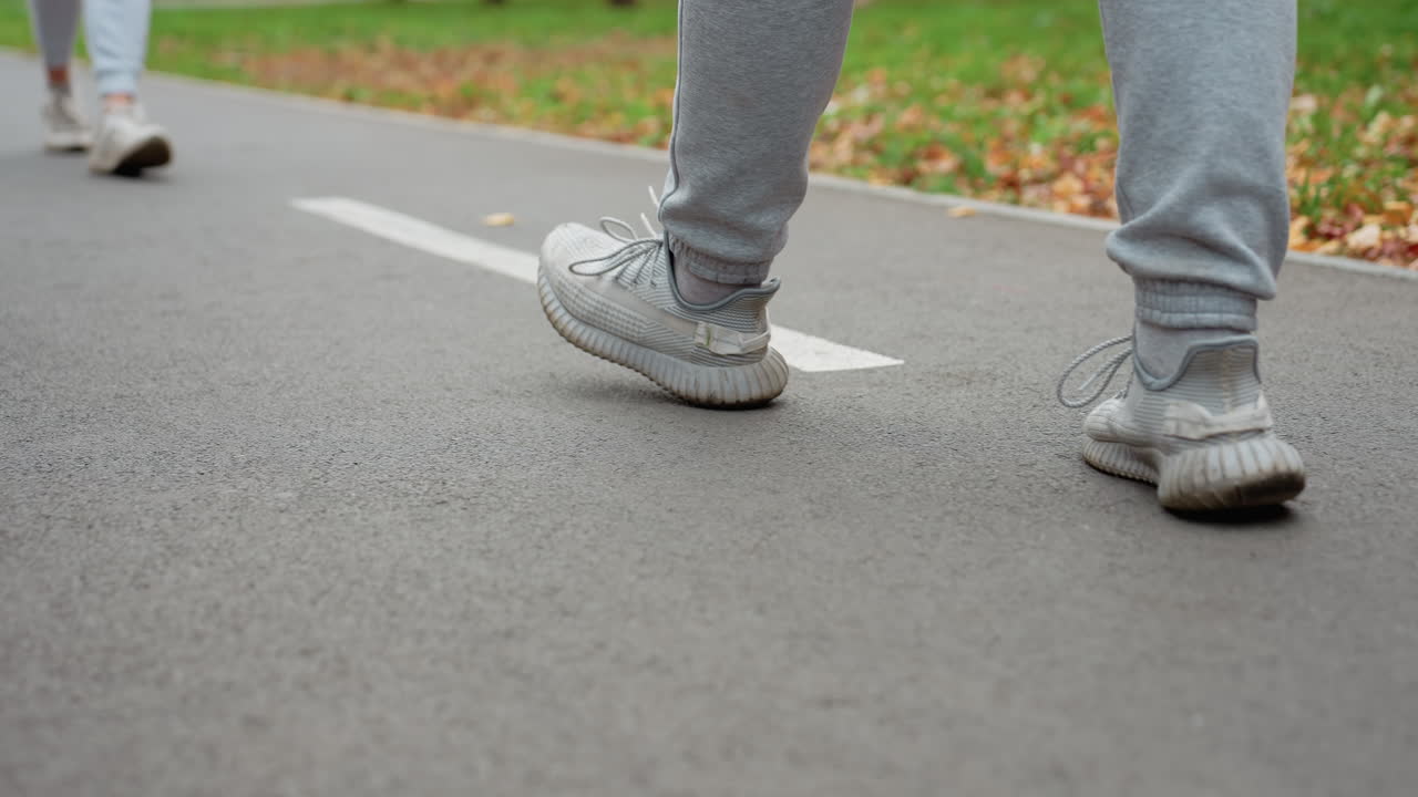 Side view of person walking on road with white markings wearing sportswear and sneakers, surrounded by greenery and scattered autumn leaves in peaceful park setting