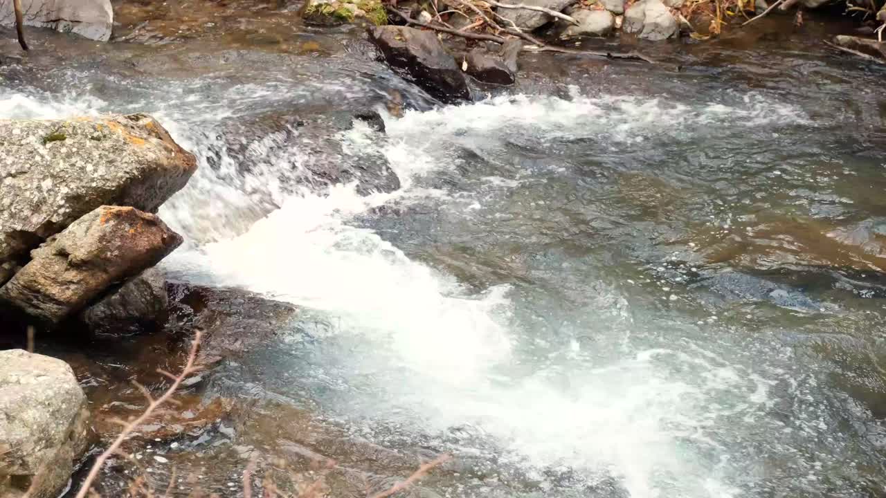 Crystal-clear water cascades over dark rocks in a mountain stream, creating white foam in a still 4K close-up view