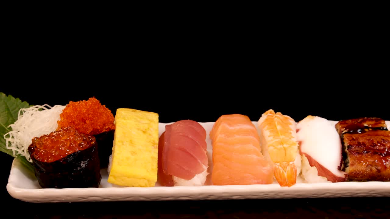 Ein rechteckiger Teller mit verschiedenen Sushi-Sorten, darunter Nigiri, Sashimi und Tamago, dreht sich sanft vor einem schwarzen Hintergrund unter weicher, gleichmäßiger Beleuchtung.