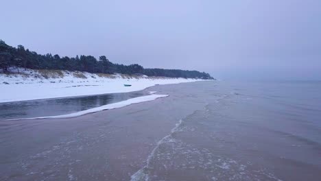 Aerial-view-of-Baltic-sea-coast-on-a-overcast-winter-day-with-green-coastal-fisherman-boat,-beach-with-white-sand-covered-by-snow,-coastal-erosion,-establishing-drone-shot-moving-forward