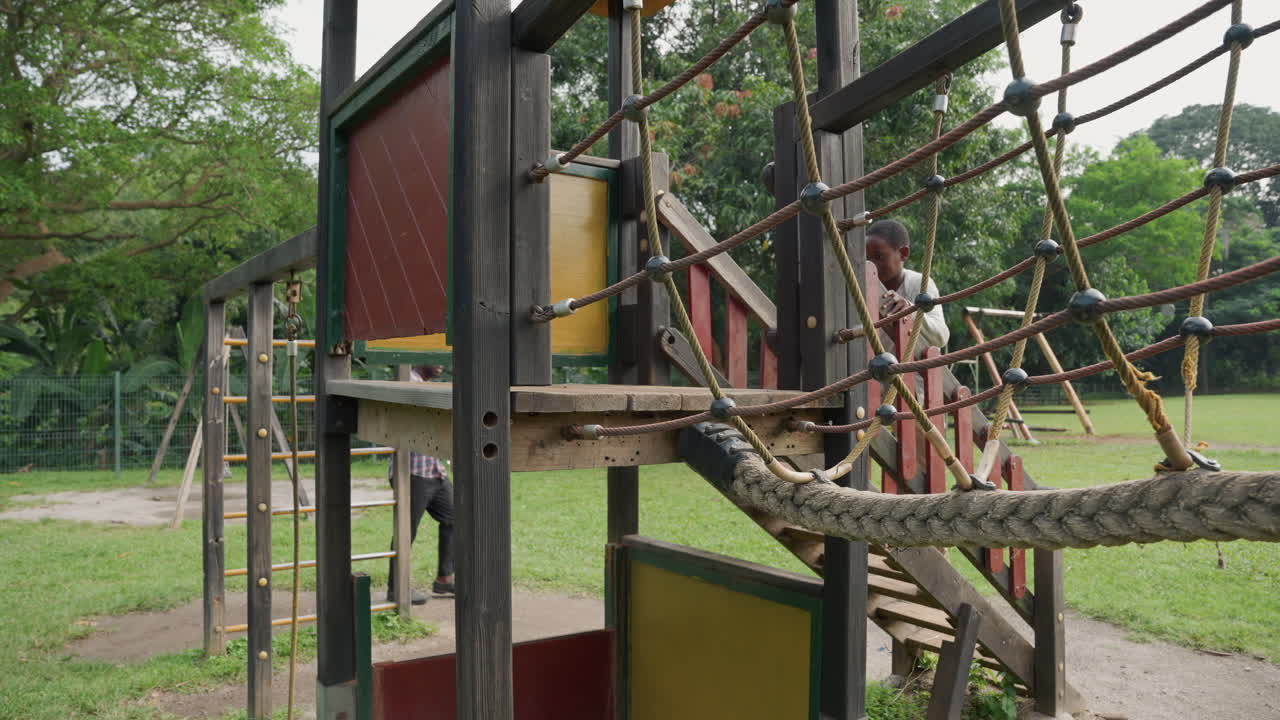 Father son climbing rope bridge wooden playground platform, green park backdrop, father standing nearby offering guidance and encouragement, child gripping ropes and stepping across, summer daylight