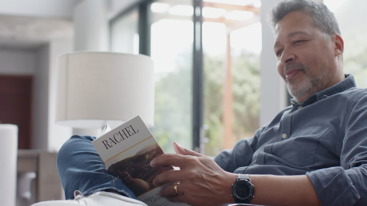 Happy senior biracial man reading book sitting in living room, slow motion