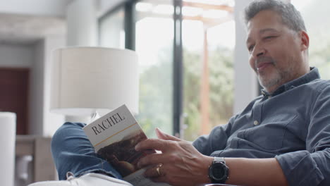 happy senior biracial man reading book sitting in living room, slow motion