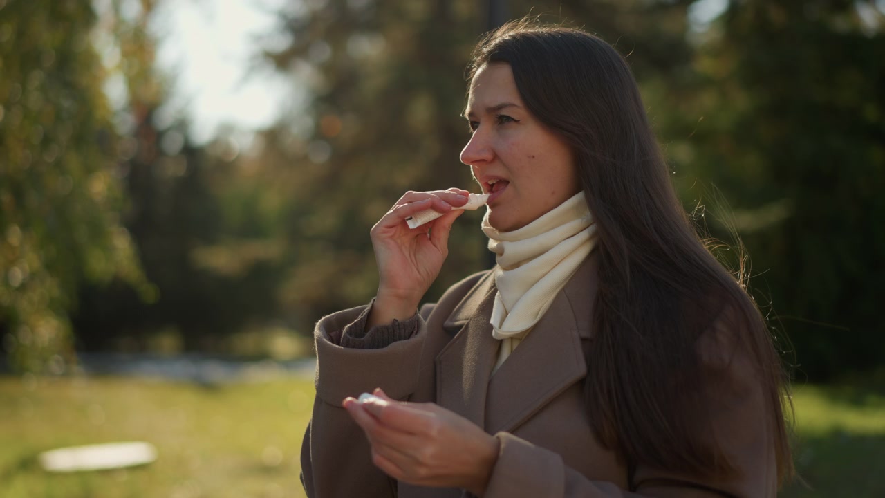 Primo piano di una donna che applica balsamo labbra all'aperto con luce naturale. La donna tiene il prodotto con una mano mentre lo usa, indossando un cappotto marrone e una sciarpa color crema, sullo sfondo di un parco verde con alberi.