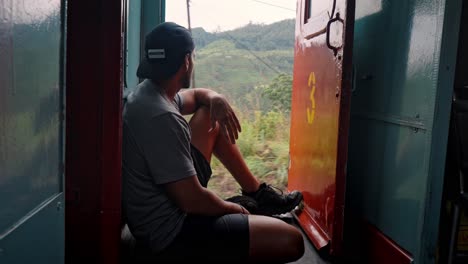 a man sits at the open door of a moving train on the scenic ella to kandy route in sri lanka, peacefully enjoying the lush landscape of tea plantations and rolling hills.