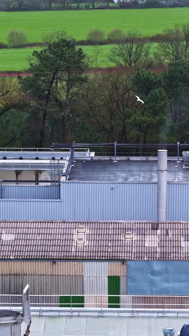 Aerial vertical tilt up from industrial factory rooftop with metal walls and ventilation to rural village in european countryside landscape with green fields
