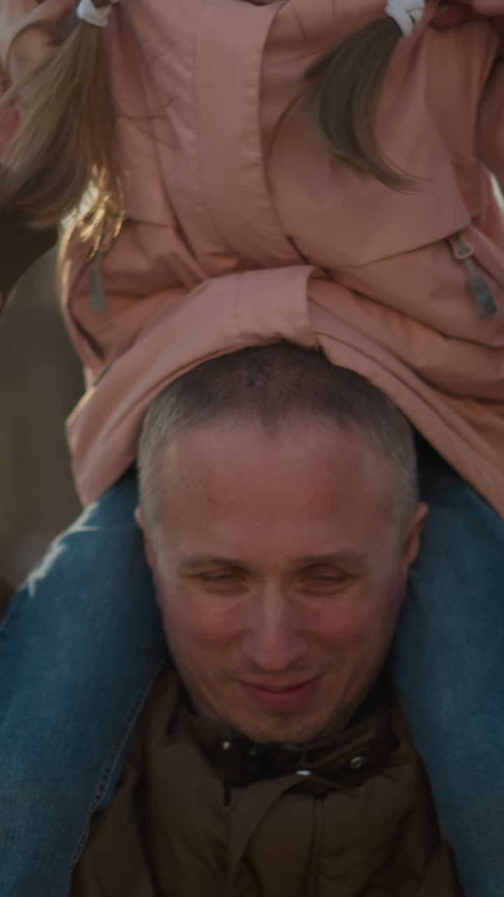 A man wearing a brown jacket carries a little girl in a pink cap and jacket on his neck, creating a joyful and playful moment outdoors in a park setting with trees and sunlight in the background