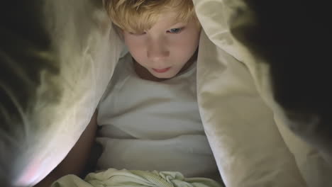 little boy using a tablet watching a movie covered by the bed sheet at night