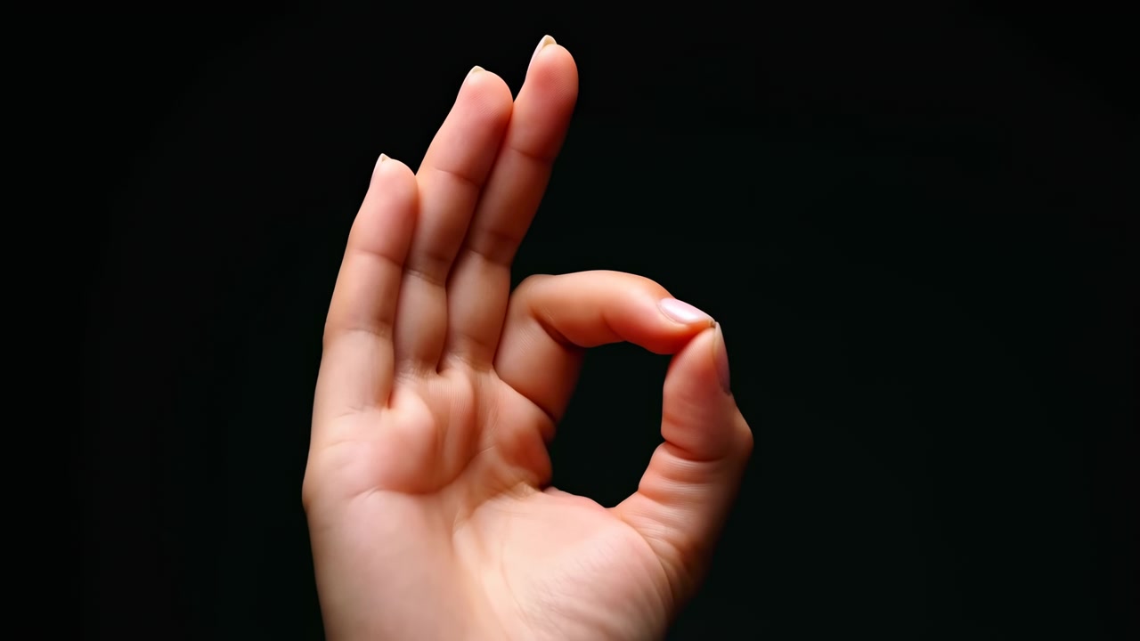 Primo piano di una mano che fa il segno dell'okay su sfondo nero, l'illuminazione da studio evidenzia la manicure curata e la pelle.