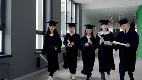 group of happy  kindergarten students in cap and gown walking in the school corridor while holding diploma and celebrating their graduation