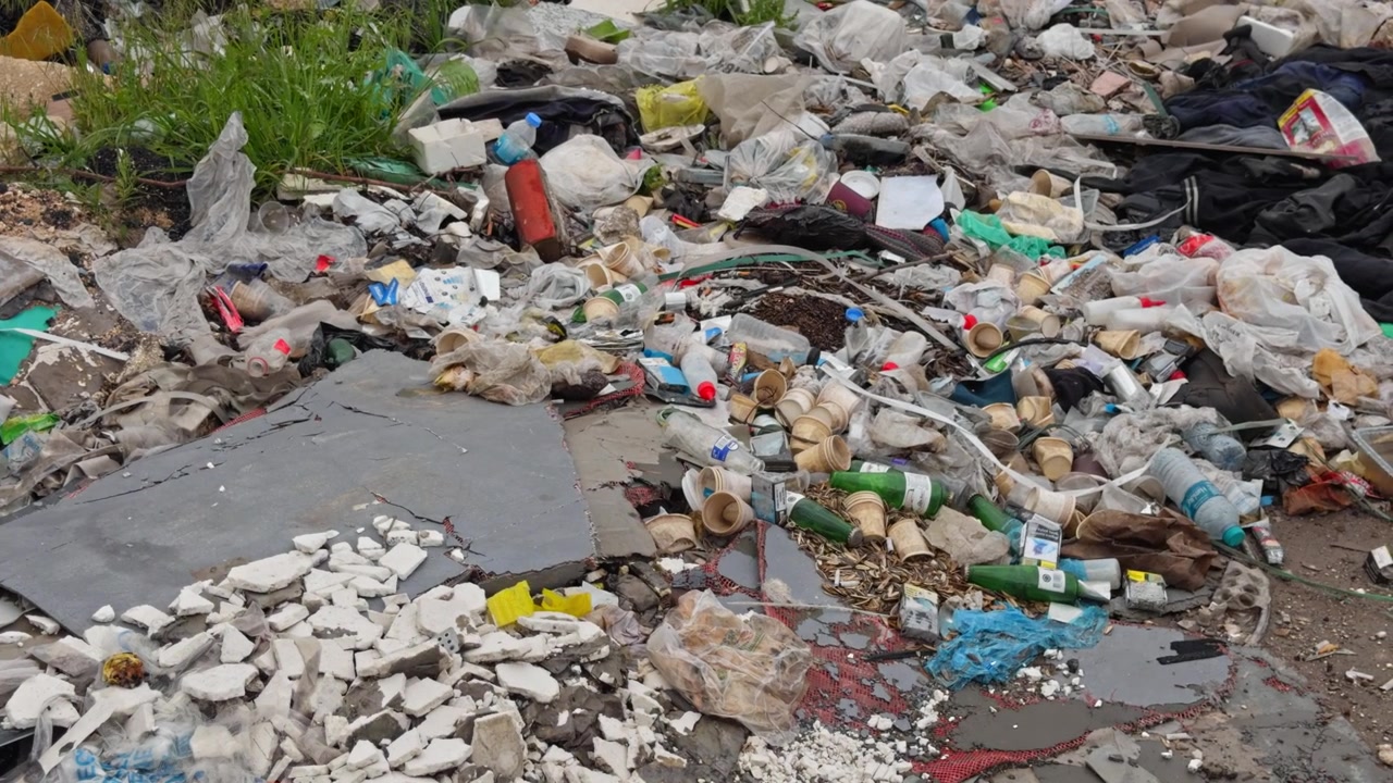 Imagens cinematográficas 4K de entulho de construção, lixo plástico e detritos mistos espalhados ao longo de uma estrada industrial urbana negligenciada. Veículo abandonado e estrutura de aço visíveis
