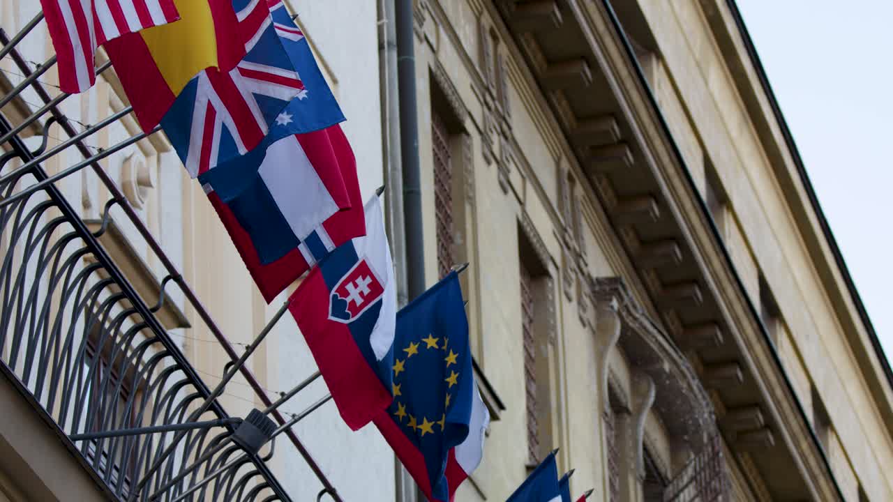 Multiple international flags wave outside ornate embassy facade, daylight, slight camera pan, old town