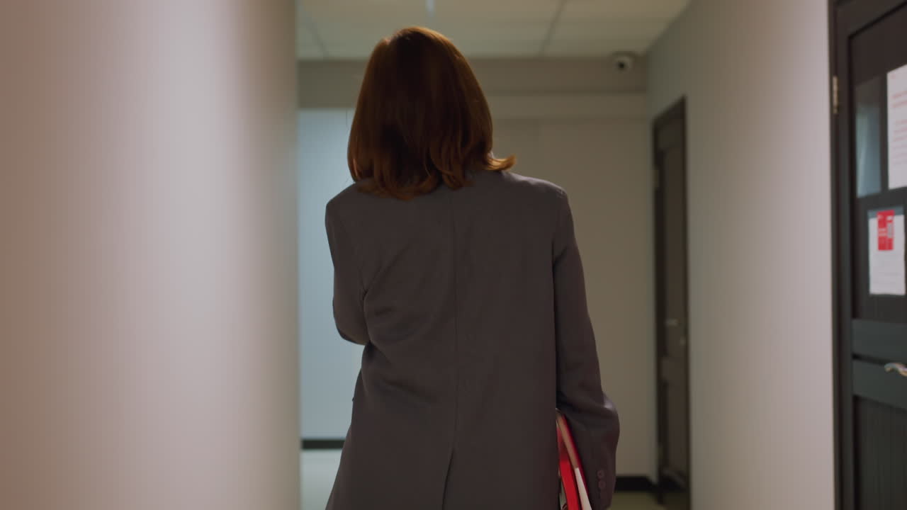 Businesswoman walking down office corridor, carrying red folder, professional environment, confident professional in gray suit, work setting, modern office, corporate workplace, career focus