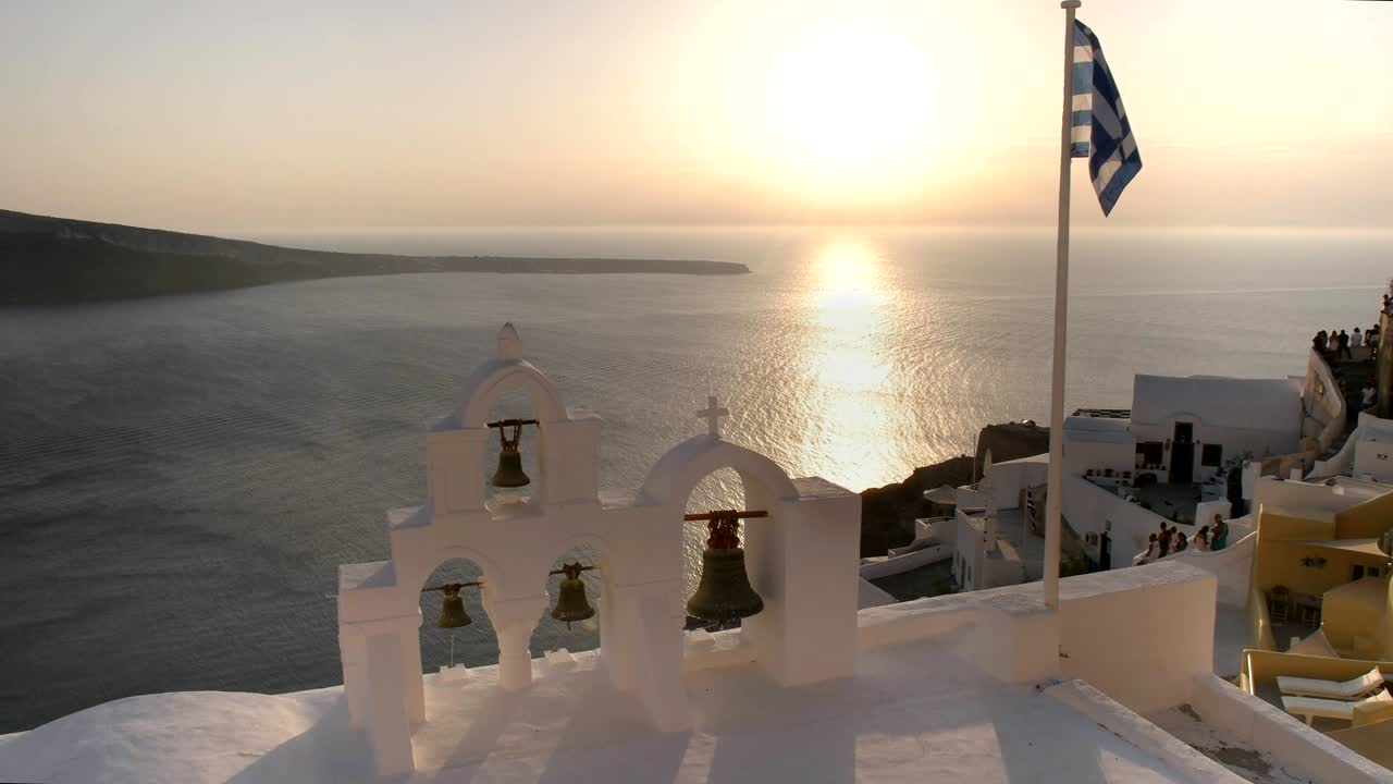 bandera griega y cuatro campanas al atardecer en oia, santorini