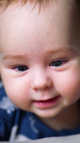 beautiful grey-eyed child in blue clothes smiles to the camera. baby boy lies on bed and moves cheerfully with his feet. close up. vertical video
