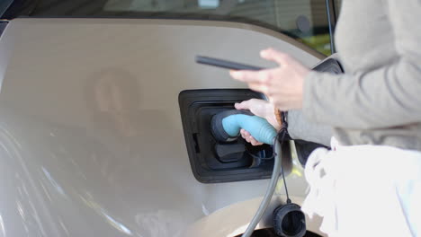 charging electric car, woman holding charging plug and smartphone in garage, copy space