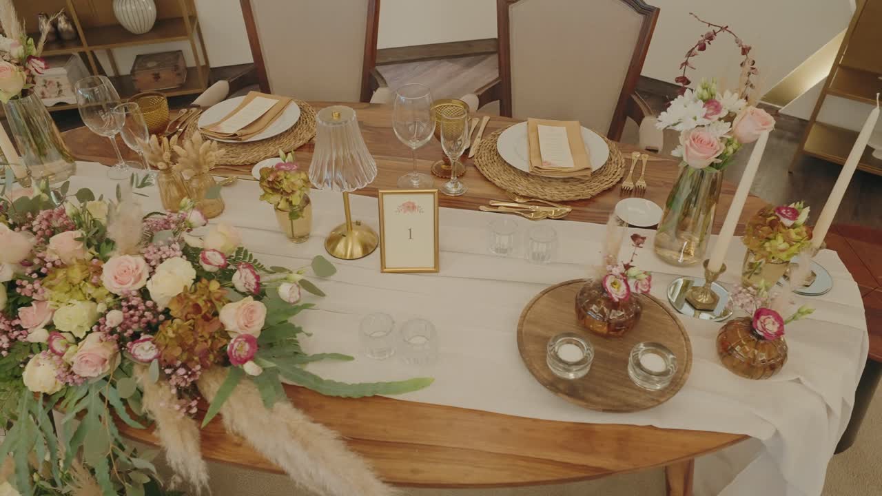 Pemandangan luas meja resepsi pernikahan yang elegan, dihiasi dengan rangkaian bunga pastel, lilin, detail emas, dan peralatan makan mewah.