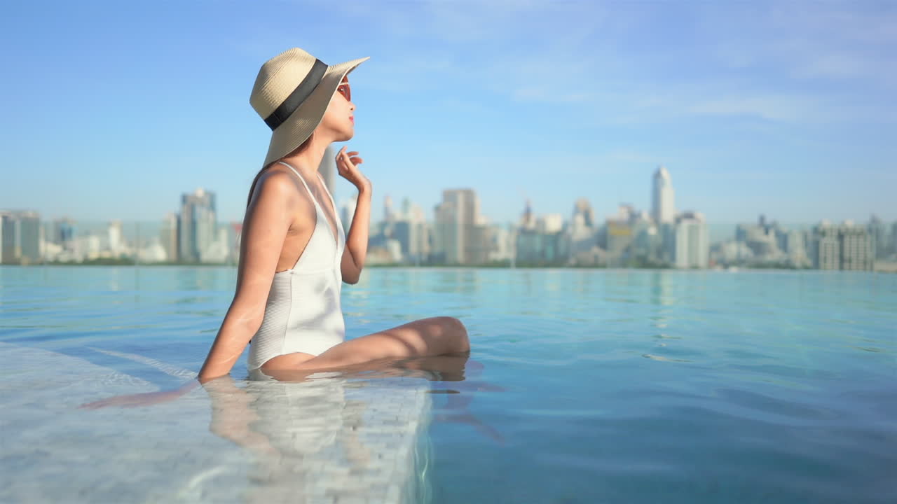 Slowmotion of young fit Asian woman sitting on the edge of the rooftop infinity pool in white swimming suit and red sunglasses on amazing blurred cityscape background
