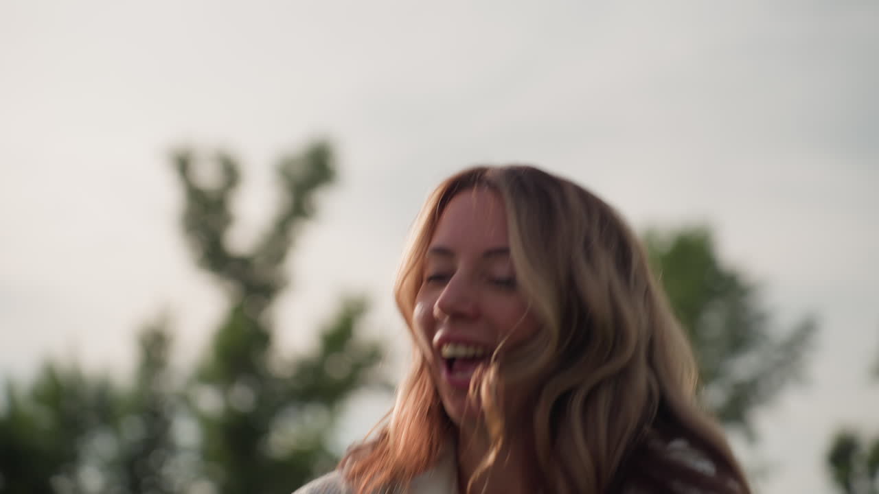 Nurturing woman spreading blanket happily in sunny park, warm sunlight through trees, breeze lifting strands, green blurred background, beaming smile, joyful family picnic preparation moment
