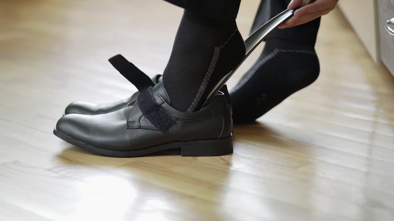 Boy is putting on new black shoes with the help of special object indoors. Classic leather shoes on velcro a male child puts on his feet with shoe spoon in the room.
