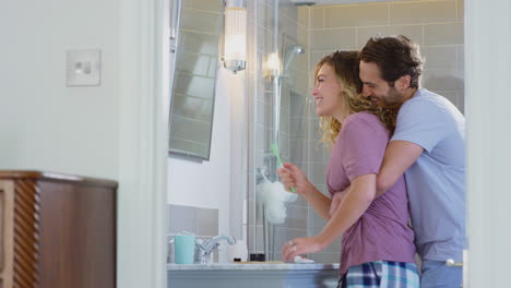 man hugging woman wearing pyjamas in en suite bathroom as she brushes her teeth