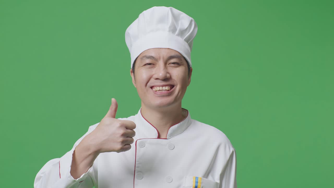 primer plano del chef asiático sonriendo y mostrando los pulgares hacia arriba gesto a la cámara mientras está de pie en el estudio de fondo de la pantalla verde