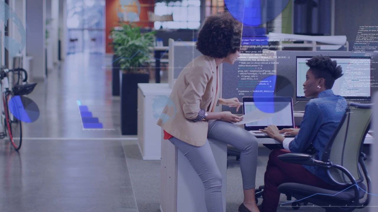 Perched colleague angling laptop and urging seated peer typing with charts animating business data