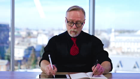 Senior man in black shirt, vest and red tie tries to write with both hands simultaneously. Man practicing handwriting on paper and I-pad. Cityscape in window at backdrop in blur.