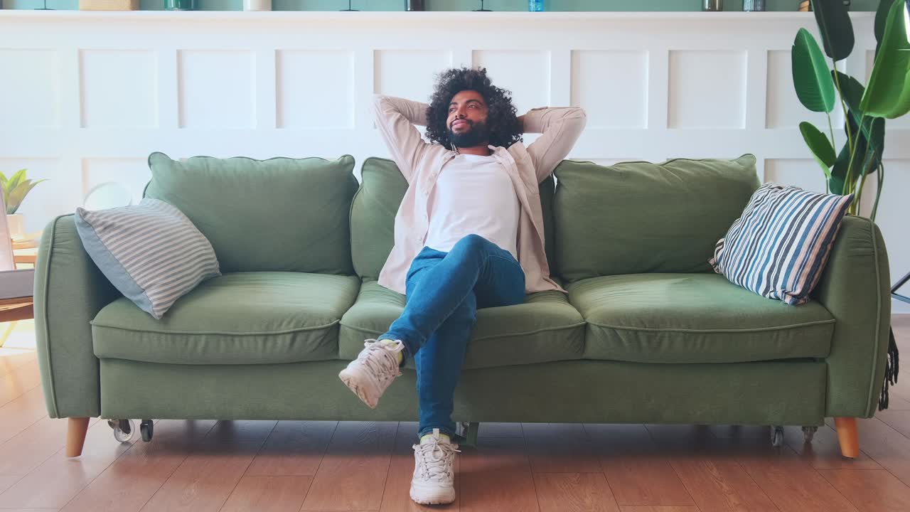 Young carefree arabian man sits on sofa leaning back and crosslegged