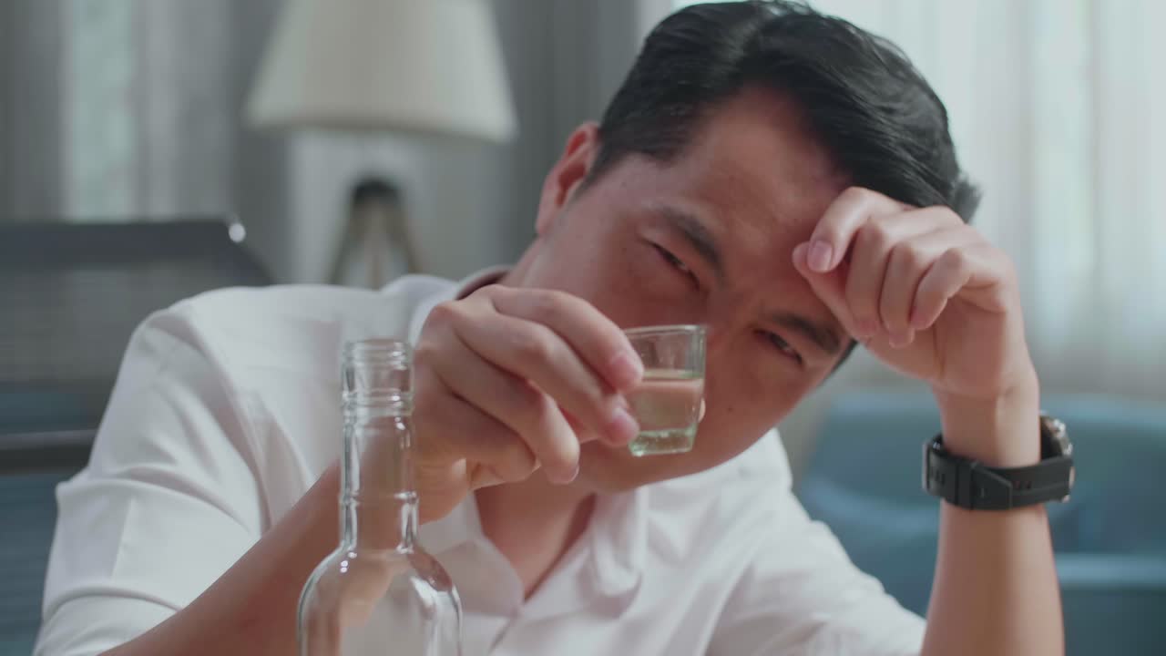 술에 취한 아시아인 남자가 집에서 술을 마시기 전에 카메라 앞에서 보드카 한 잔을 즐기고 있다.